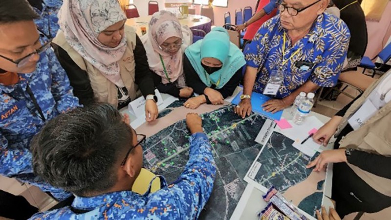 People stand grouped around a table, working together on mapping safer locations for disaster relief and emergency rescue